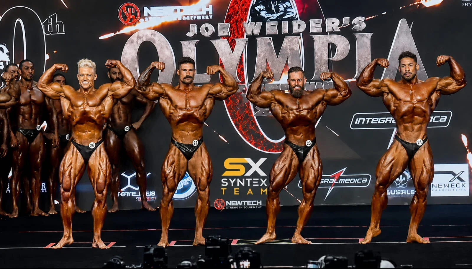Line of athletes on the Olympia stage with Syntex Team identity on the backdrop